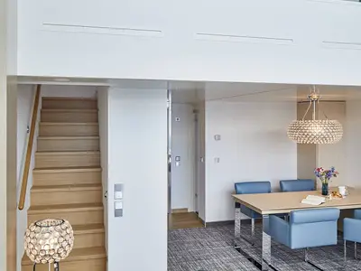 Einblick in eine moderne Penthouse Suite der AIDAnova mit Essbereich, hellem Holztisch, blauen Stühlen und Kristallleuchter. Eine Holztreppe führt nach oben. Die Suite ist elegant und hell gestaltet.
