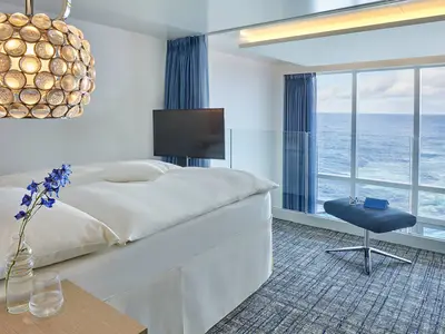 Blick in eine helle, moderne Penthouse Suite der AIDAnova mit Doppelbett, Design-Lampe, TV und einem Hocker vor der grossen Fensterfront mit weitem Meerblick.