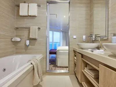 Modernes Bad einer Premium Suite an Bord der AIDAnova mit Badewanne, zwei Waschbecken, Spiegel und Meerblick durch die Suite.
