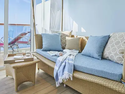 Blick in eine helle Suite an Bord der AIDAnova mit einem geflochtenen Sofa, blauen Kissen und einem Tablett mit Getränken. Im Hintergrund ist der Balkon mit Liegestuhl und Meer zu sehen.