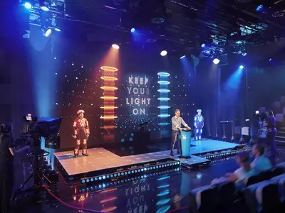 Eine hell erleuchtete Buehne im StudioX an Bord der AIDAnova zeigt einen Moderator und zwei Performer in leuchtenden Anzuegen, umgeben von Kameras und einem Publikum.