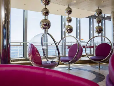 Lounge der AIDAperla mit Meerblick durch Panoramafenster. Moderne Einrichtung mit glänzenden Kugelleuchten und transparenten Schaukelstühlen mit magenta Kissen. Ein Smartphone auf einem Sessel.