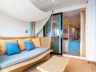 Ein heller Lanai-Balkon mit einem Rattan-Daybed, blauen und goldenen Kissen. Durch eine Glasschiebetür blickt man in die moderne Kabine der AIDAperla.