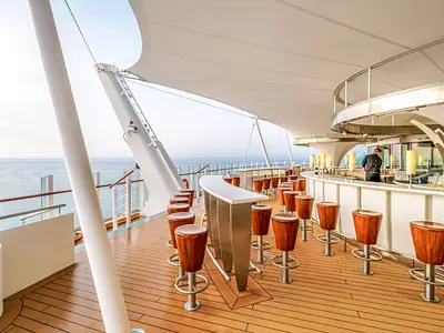 Moderne Aussenbar der AIDAperla. Ein Barkeeper mixt Getraenke. Weisser Tresen, Holzbarhocker, Meerblick und Segeldach.