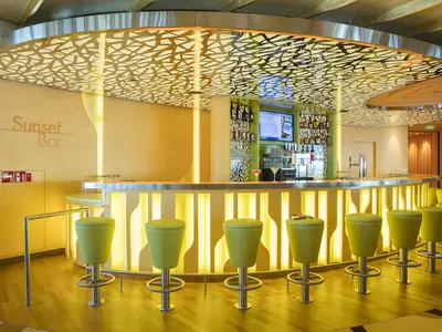 Die moderne Sunset Bar an Bord der AIDAperla ist in warmen Gelbtönen gehalten. Eine beleuchtete Theke mit grünen Barhockern dominiert den Raum. Die Decke zeigt ein auffälliges, beleuchtetes Muster. Hinter der Theke sind Flaschenregale und ein Bildschirm sichtbar.