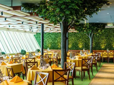 Das geräumige Markt Restaurant an Bord der AIDAperla ist hell und freundlich eingerichtet. Zahlreiche gelb gedeckte Tische und Holstühle stehen zwischen dekorativen Bäumen. Eine grüne Pflanzenwand und große Fenster sorgen für eine frische, natürliche und lichtdurchflutete Atmosphäre.