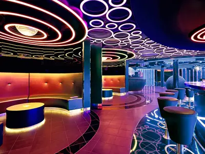 Die futuristische Diskothek D6 an Bord der AIDAperla bei Nacht. Auffällige Leuchtkreisen an Decke und Wänden, moderne Sitznischen und eine Bar mit Hockern in leuchtenden Farben.