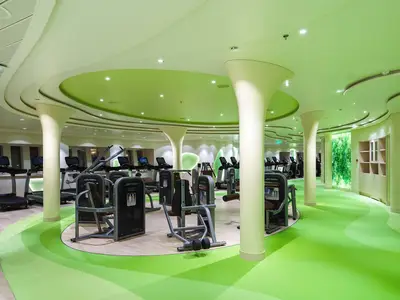 Innenansicht des Body & Soul Sportbereichs an Bord der AIDAperla. Zahlreiche moderne Life Fitness Geräte stehen auf einem hellen Holzboden, umgeben von einem hell- und dunkelgrünen, organisch geformten Boden. Helle Säulen und eine grüne Decke mit eingebauten Lichtern prägen den Raum. Im Hintergrund ist eine leuchtende Wand mit Pflanzenmotiv zu sehen. Keine Personen.