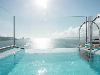 Infinity Pool an Bord der AIDAperla mit Meerblick auf Küste und Hafen. Sonne glitzert auf dem Wasser. Gläserne Brüstung, Poolleiter. Keine Personen.