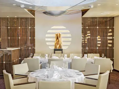 Das elegante Rossini Restaurant an Bord der AIDAperla ist festlich gedeckt. Helle Stühle und weiße Tischdecken sind sichtbar. Eine goldene Skulptur vor einer runden Lichtinstallation prägt den Raum.