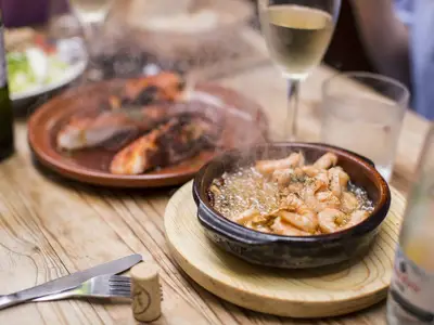 Nahaufnahme von dampfenden Gambas al Ajillo und weiteren Tapas auf einem Holztisch. Ein Glas Weißwein steht bereit im Restaurant der AIDAperla.