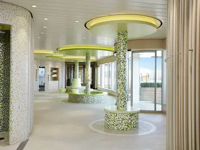 Ein heller, moderner Spa-Bereich an Bord der AIDAperla mit grünen Mosaik-Duschen, runden Säulen und Sitzgelegenheiten. Große Fenster bieten Blick auf das Außendeck.