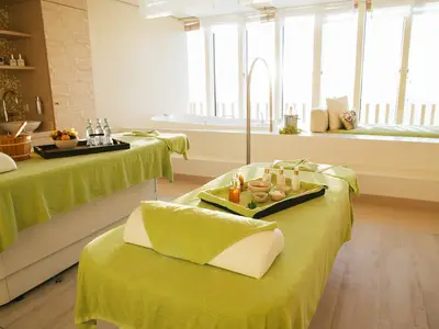 Ein heller Wellnessbereich an Bord der AIDAperla mit zwei grünen Massageliegen und verschiedenen Spa-Produkten. Im Hintergrund sind eine Badewanne und große Fenster mit Blick nach draußen zu sehen.