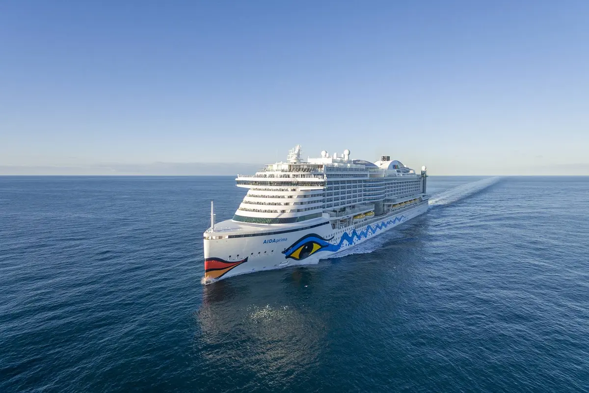 AIDAprima, ein modernes Kreuzfahrtschiff, fährt auf blauem Meer unter klarem Himmel. Das weisse Schiff mit dem bunten Kussmund am Bug zieht eine lange Heckwelle.