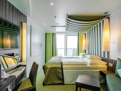 Innenansicht einer modernen Panoramakabine an Bord der AIDAprima. Ein Doppelbett, ein Sofa mit Tisch, TV und ein Schreibtisch sind zu sehen. Die Kabine hat große Fenster mit Zugang zu einem Balkon.