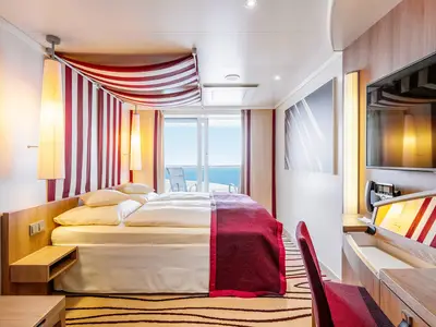 Moderne Verandakabine an Bord der AIDAprima mit Doppelbett, Schreibtisch, TV und Balkon. Das Dekor ist rot-weiß gestreift, der Blick geht auf das Meer.