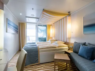 Blick in eine moderne Balkonkabine an Bord der AIDAprima mit Doppelbett, Sofa, Beistelltisch und Zugang zu einem Balkon. Warme Beleuchtung.