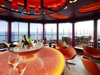 Blick in die moderne Vinothek an Bord der AIDAprima mit Champagner auf Eis, orangenen Sesseln und Meerblick durch große Fenster.