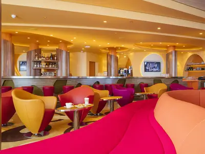 Moderne, farbenfrohe Bar-Lounge 'Café Mare' an Bord der AIDAprima mit runden Barhockern, Sesseln in Rot, Gelb und Pink, sowie einer leuchtenden Decke.