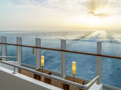 Blick von der AIDAprima Lanai Bar bei Sonnenuntergang über das weite Meer, mit der Schiffspur und modernen Stehlampen auf Holztischen. Das Glasgeländer und der Handlauf rahmen die Szene ein.