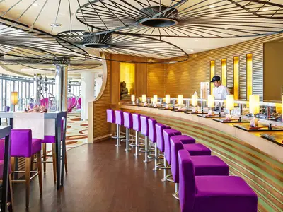 Eine stilvolle Sushi Bar an Bord der AIDAprima mit einem langen Tresen, purpurfarbenen Barhockern und einem Koch hinter Glas. Im Hintergrund sind weitere Tische und große Fenster zu sehen.