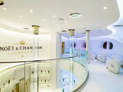 Innenansicht der Moët & Chandon Spray Bar an Bord der AIDAprima. Futuristische weiße Möbel, beleuchtete Bläulichtsäulen und eine geschwungene Glasbalustrade sind zu sehen. Die Moët & Chandon-Logowand dominiert den Hintergrund.