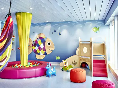 Ein lebhaft gestalteter Kinderbereich mit Ballbecken, Rutsche und Klettergerüst an Bord der AIDAprima. Die Wand ist mit einem Unterwasser-Motiv und großen bunten Fischen dekoriert. Das Tageslicht strömt durch große Fenster.