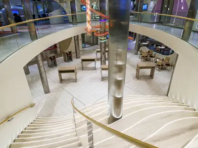 Modernes Atrium der AIDAprima. Eine beleuchtete Treppe führt zu Sitzbereichen mit Tischen und Stühlen auf hellem Fliesenboden. Farbige Lichtinstallation umgibt Säule.