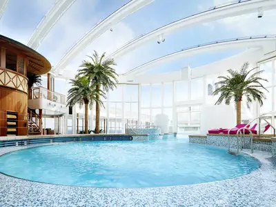 Der Beach Club an Bord der AIDAprima zeigt einen großen Pool mit türkisfarbenem Wasser, umgeben von Liegen, künstlichen Palmen und einer Holzstruktur unter einem verstellbaren Glasdach.