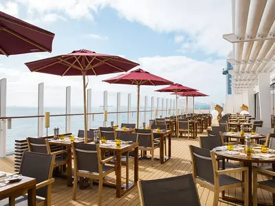 Der Außenbereich eines Restaurants an Bord der AIDAprima. Gedeckte Tische und Stühle unter roten Sonnenschirmen auf einem Holzdeck, mit weitem Meerblick.