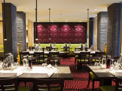 Das East Restaurant an Bord der AIDAprima zeigt mehrere gedeckte Tische, stilvolle Einrichtung mit Stein- und Bambuselementen und eine rote Wanddekoration. Der Raum ist hell beleuchtet, ohne Personen.