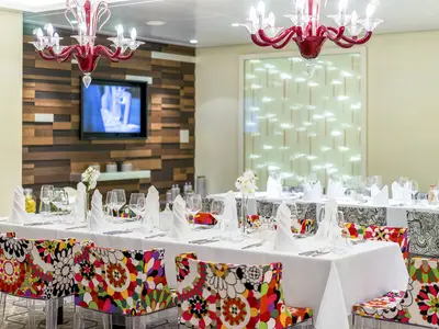 Ein modernes Restaurant an Bord der AIDAprima mit festlich gedeckten Tischen und bunten, floralen Stühlen. Rote Kronleuchter und eine Holzwand mit Fernseher im Hintergrund.