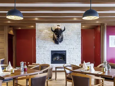 Einladendes Restaurant an Bord der AIDAprima. Die Szene zeigt gedeckte Tische, rote Wände, einen Steinkamin mit Bisonkopf-Dekoration und Holzbalken an der Decke.