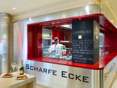 Ein lächelnder Koch serviert Currywurst an der Scharfen Ecke an Bord der AIDAprima. Die moderne Theke ist rot und silberfarben, mit Preistafeln und Champagnerflaschen.