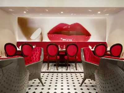 Modernes Restaurant 'French Kiss' an Bord der AIDAprima mit glänzend roten Lippen als Wandbild, rotem Samtgestühl und Schwarz-Weiß-Fliesen. Tische sind für Gäste gedeckt.