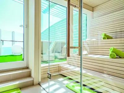 Blick in eine moderne Sauna an Bord der AIDAprima. Die Holzbänke sind mit gerollten grünen Handtüchern dekoriert. Durch eine Glaswand blickt man auf einen Balkon mit Kunstrasen und Meerblick.
