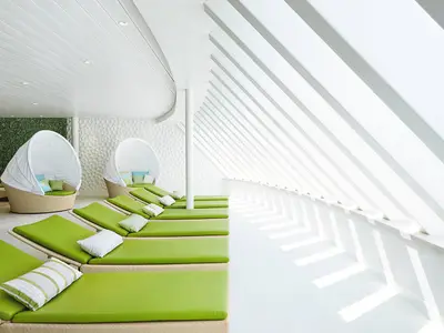 Blick in den Body & Soul Organic Spa an Bord der AIDAprima. Helle Liegen mit gruenen Auflagen und ueberdachte Rattan-Sessel laden zum Entspannen ein. Sonnenlicht faellt durch Lamellenfenster. Ohne Personen.