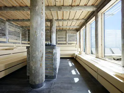 Kelosauna an Bord der AIDAprima mit naturbelassenen Holzstämmen und hellen Bänken. Durch die Fensterfront blickt man auf ein sonniges Außendeck mit Liegestühlen.