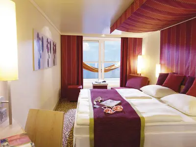 Moderne Balkonkabine an Bord der AIDAsol mit Doppelbett, violetter Decke und roten Kissen. Eine Hängematte hängt vor dem Balkon, dahinter blauer Himmel.