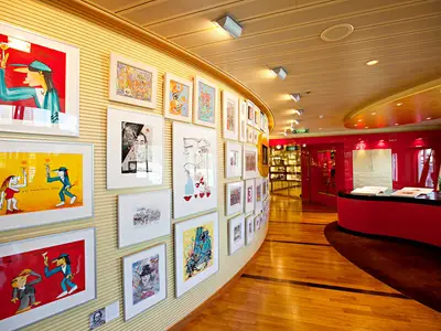 Ein heller Flur an Bord der AIDAsol, dessen geschwungene Wand mit vielen gerahmten, bunten Kunstwerken behängt ist. Im Hintergrund eine leuchtend rote Rezeption.