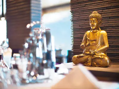 Eine goldene Buddha-Statue, friedlich meditierend, ist im East Restaurant der AIDAsol zu sehen. Sie steht auf einem dunklen Tresen vor einer Holzvertäfelung, mit Tageslicht im Hintergrund.