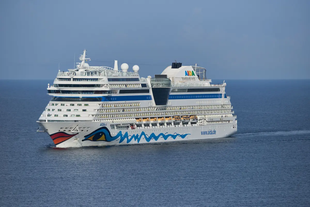 Die AIDAstella fährt von links nach rechts auf dem weiten blauen Meer. Der weisse Rumpf mit dem farbigen Augen- und Kussmund-Logo ist gut zu erkennen, ebenso die blauen Streifen und die Decksaufbauten unter blauem Himmel.