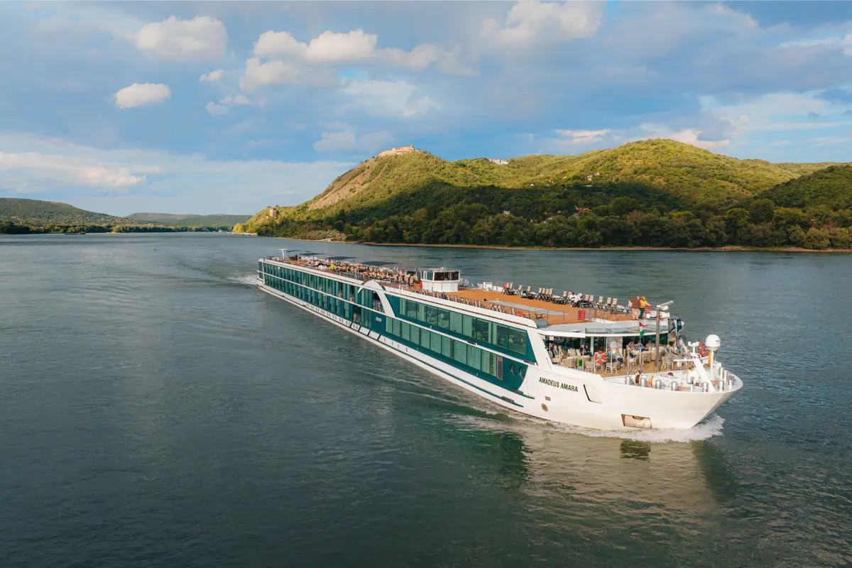 Der Flusskreuzer Amadeus Amara gleitet bei Tageslicht auf einem breiten Fluss. Grüne, bewaldete Hügel säumen die Ufer unter einem teilweise bewölkten Himmel. Das Schiff hat mehrere Decks mit Passagieren auf dem Sonnendeck.
