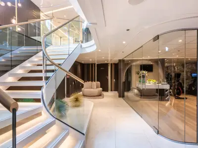 Eine helle, moderne Atrium-Lounge auf der Amadeus Amara. Links eine geschwungene weisse Treppe mit Glasgeländer. Rechts ein Fitnessstudio hinter Glas, davor eine Sitzecke und Deko-Elemente.