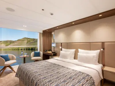 Helle, moderne Kabine auf dem Mozart and Strauss Deck der Amadeus Amara mit Doppelbett, einem kleinen Tisch mit Snacks und zwei Sesseln vor einem grossen Panoramafenster. Draussen sind ein Fluss und bewaldete, terrassierte Weinberge zu sehen.