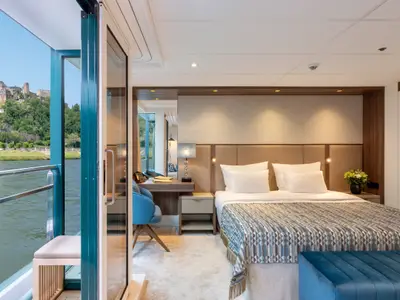 Eine elegant eingerichtete, helle Suite auf der Amadeus Amara mit einem gemusterten Doppelbett, einem Schreibtisch und einer Glasfront zu einem Fluss mit einer Burg im Hintergrund.