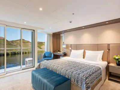 Eine helle, moderne Suite auf der AMADEUS Amara. Sie zeigt ein grosses Bett, eine Sitzbank, einen Schreibtisch und eine Glasfront mit Blick auf einen Fluss und begrünte Hügel mit Weinbergen. 