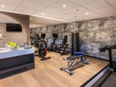 Ein gut ausgestatteter Fitnessbereich auf der Amadeus Amara mit hellem Holzboden, einem Laufband, zwei Ergometern, einer Hantelbank und einem Hantelständer. 