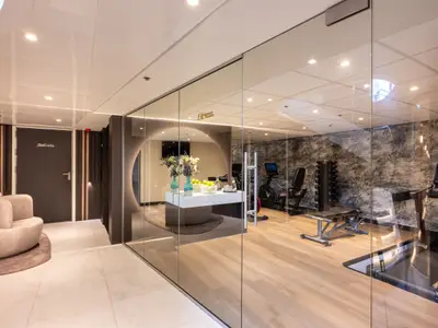 Modernes Fitnessstudio auf der Amadeus Amara, hinter Glaswand. Laufbänder, Hanteln, Fahrräder, Tisch mit Obst/Wasser. Davor: Lounge-Sitzgruppe und Tür mit "Staff only".