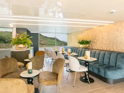 Cafe Vienna: Elegante Lounge mit tealblauer Sitzbank, goldenen Sesseln, kleinen Tischen und großen Fenstern zum Fluss und grünen Weinbergen.